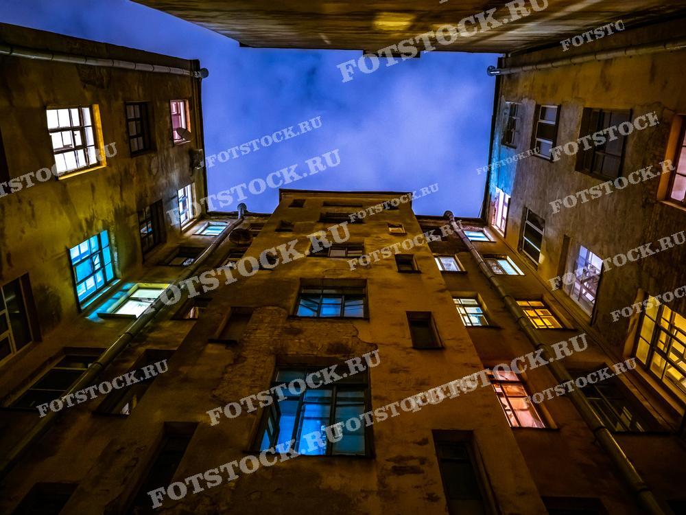 На фотографии изображено небо, видимое из двора-колодца в Санкт-Петербурге ночью. Снимок выделяется контрастом подсвеченных окон и темных стен зданий. Это изображение подойдет для иллюстраций архитектурных особенностей Санкт-Петербурга, блогов о городской жизни и исторических исследований.